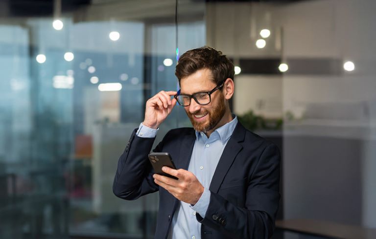 Moški s pametnim telefonom stoji pred stekleno pisarno in si popravlja očala. Oblečen je v temno modro obleko in svetlo modro srajco.
