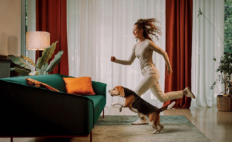 Dekle v bež oblačilih in kužek tečeta k zelenemu kavču z oranžnimi blazinami. V prostoru so luč, velike rastline in zavese.