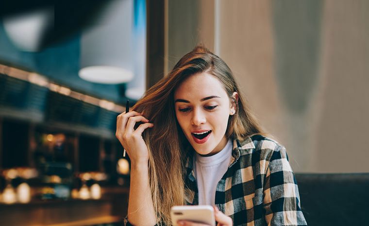 Dekle z dolgimi lasmi in v karirasti srajci sedi v notranjosti kavarne in gleda v telefon. V ozadju so vidne luči in notranja oprema kavarne.