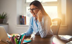 Dekle z očali sedi za mizo v domači pisarni, govori po telefonu in dela na prenosniku.