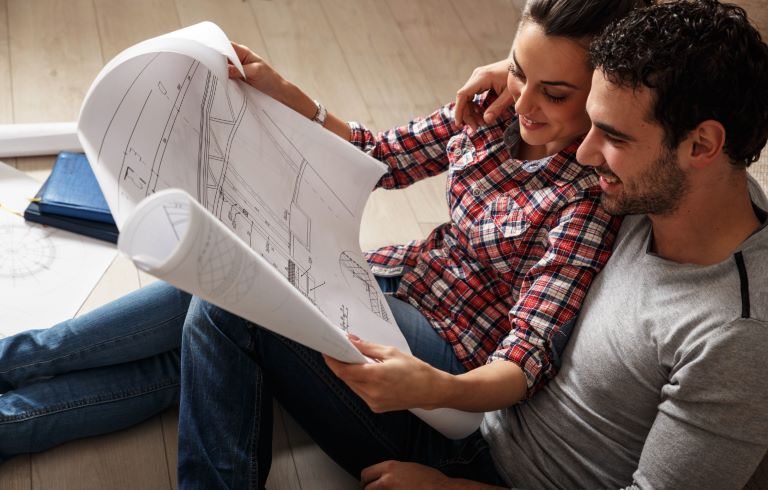 Par sedi na tleh in si ogledujeta arhitekturni načrt. Načrt vsebuje tehnične risbe in mere. Ženska nosi karirasto srajco, moški pa sivo majico.