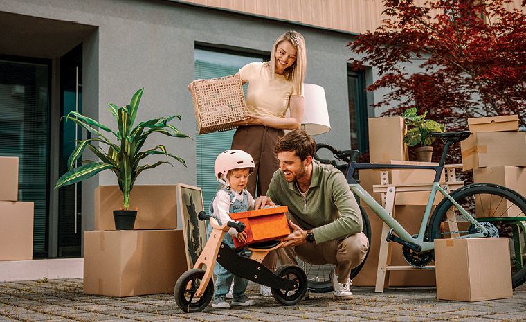 Družina se seli v nov dom. Stojijo pred hišo, obdani so s škatlami. Otrok in oče z oranžno škatlo čepita ob poganjalčku, mama stoji za njima.