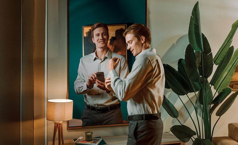 Dva moška stojita pred ogledalom. Levi drži telefon in gleda v ogledalo, desni si zapenja gumbe na srajci. V ozadju se vidi svetilka in velika zelena rastlina.