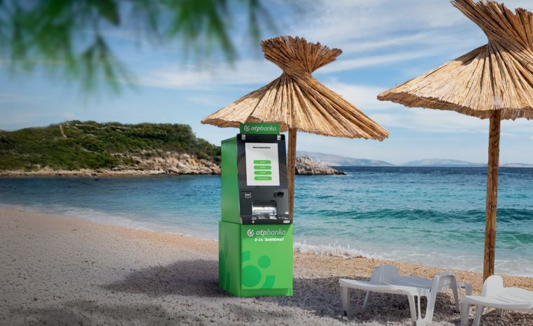Dva slamnata senčnika in bela ležalnika na plaži ob morju. Ob senčniku stoji zelen bankomat z logotipom OTP banke. Dva slamnata senčnika in bela ležalnika na plaži ob morju. Ob senčniku stoji zelen bankomat z logotipom OTP banke.