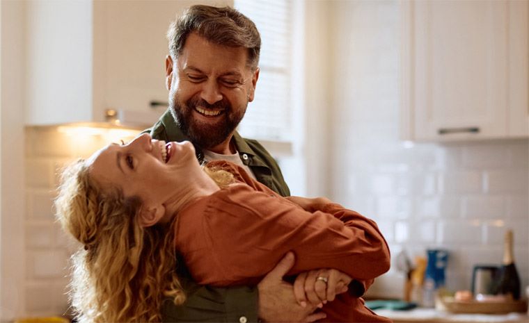 Fotografija para, kjer moški drži žensko v naročju v svetli kuhinji, pri čemer prizor deluje topel in skrben.