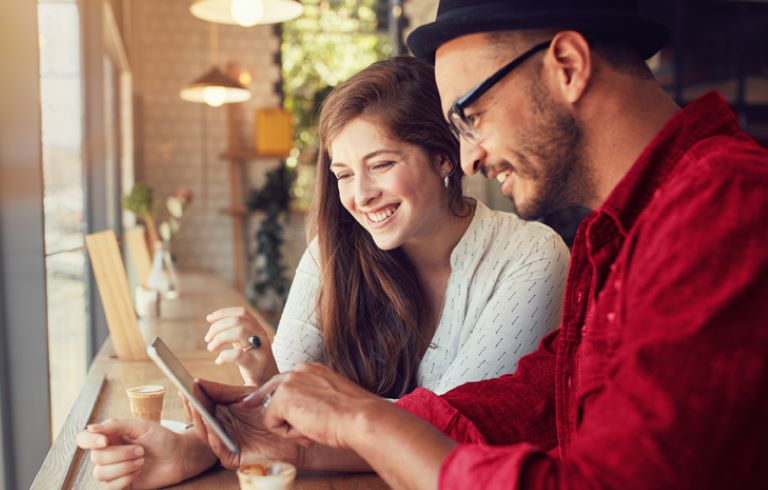 Moški v rdeči in ženska v beli srajci sedita ob oknu v kavarni. Gledata v zaslon pametnega telefona. Na mizi je skodelica kave.