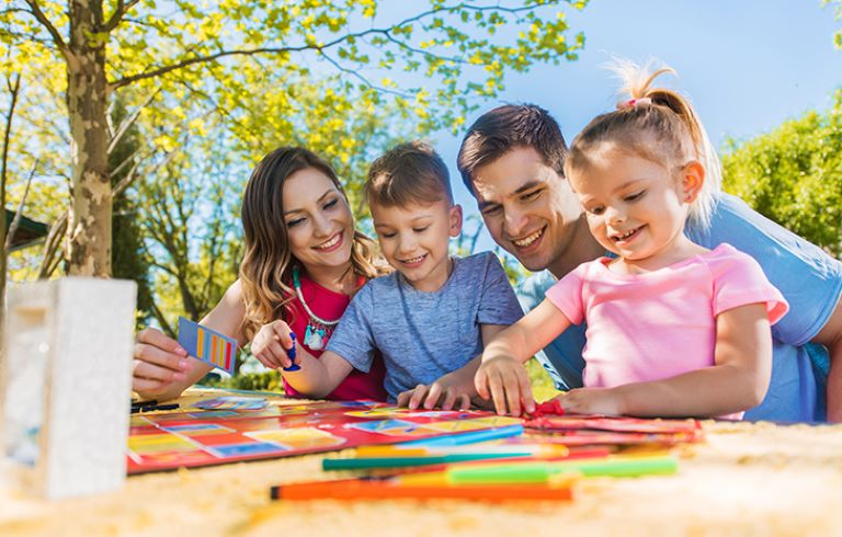 Družina na sončen dan sedi za mizo na prostem in ustvarja z barvnimi papirji, flomastri in škarjami. Družina na sončen dan sedi za mizo na prostem in ustvarja z barvnimi papirji, flomastri in škarjami.