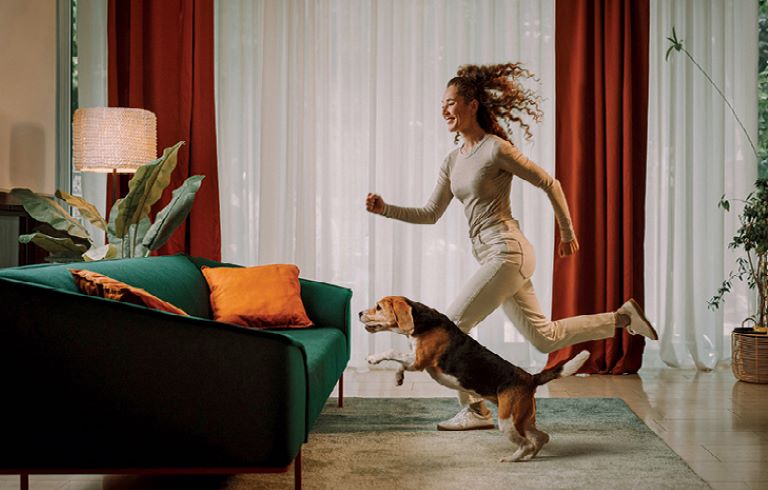 Dekle v bež oblačilih in kužek tečeta k zelenemu kavču z oranžnimi blazinami. V prostoru so luč, velike rastline in zavese.