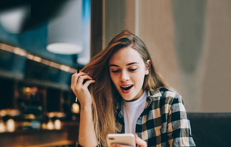 Dekle z dolgimi lasmi in v karirasti srajci sedi v notranjosti kavarne in gleda v telefon. V ozadju so vidne luči in notranja oprema kavarne.