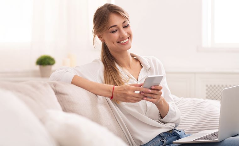 Mlada ženska sedi na kavču in gleda v pametni telefon. Oblečena je v belo srajco, ima rjave lase, ki so speti v čop. V ozadju je okno z zavesami ter majhna zelena rastlina, ki stoji na polici.