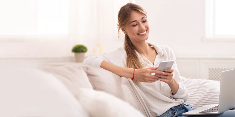 Mlada ženska sedi na kavču in gleda v pametni telefon. Oblečena je v belo srajco, ima rjave lase, ki so speti v čop. V ozadju je okno z zavesami ter majhna zelena rastlina, ki stoji na polici.