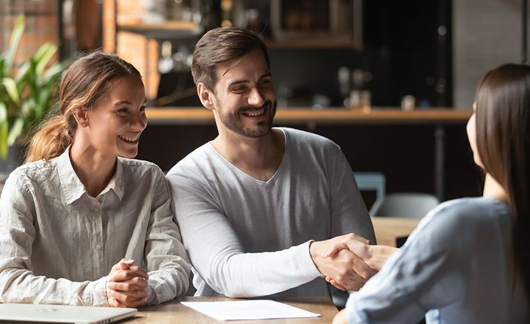 Moški in dve ženski sedijo na poslovnem sestanku za mizo v pisarni. Moški se rokuje z žensko, ki mu sedi nasproti.