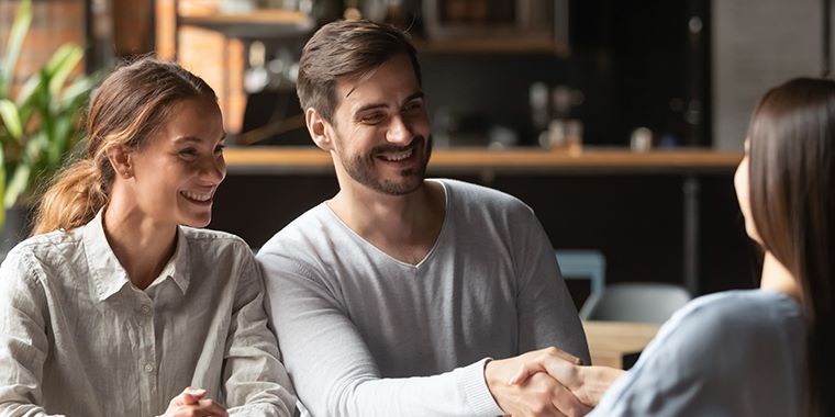 Moški in dve ženski sedijo na poslovnem sestanku za mizo v pisarni. Moški se rokuje z žensko, ki mu sedi nasproti.