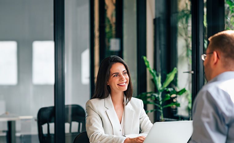 Moški in ženska sedita na sestanku v pisarni. Ženska nosi bel suknjič in ima črne lase, moški je v svetlo modri srajci.