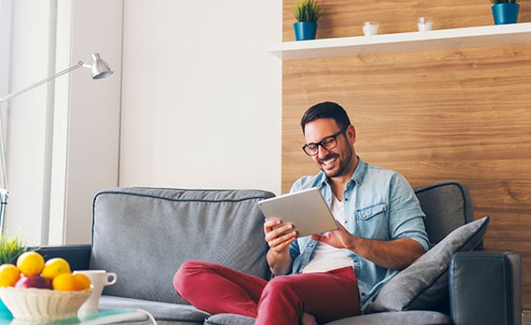 Moški sedi na sivem kavču in gleda v tablico. V ozadju je lesena stena. Levo stoji svetilka, na mizi pred kavčem pa skleda s sadjem.