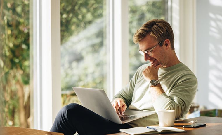 Moški sedi za mizo ob velikem oknu in dela na prenosnem računalniku. Na mizi so skodelica kave, zvezek in pisalo.
