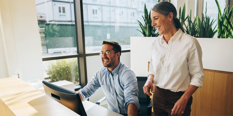 Moški v modri srajci dela na računalniku, ob njem stoji ženska v beli srajci. V ozadju rastline, velika okna, skozi katera se vidi stavba.