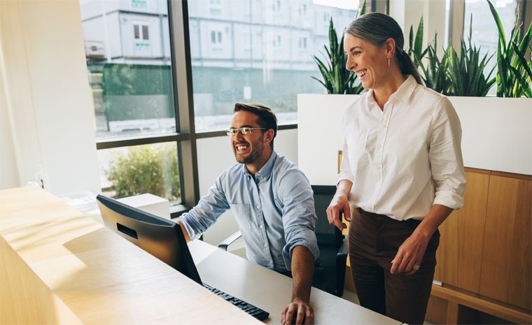 Moški v modri srajci dela na računalniku, ob njem stoji ženska v beli srajci. V ozadju rastline, velika okna, skozi katera se vidi stavba.