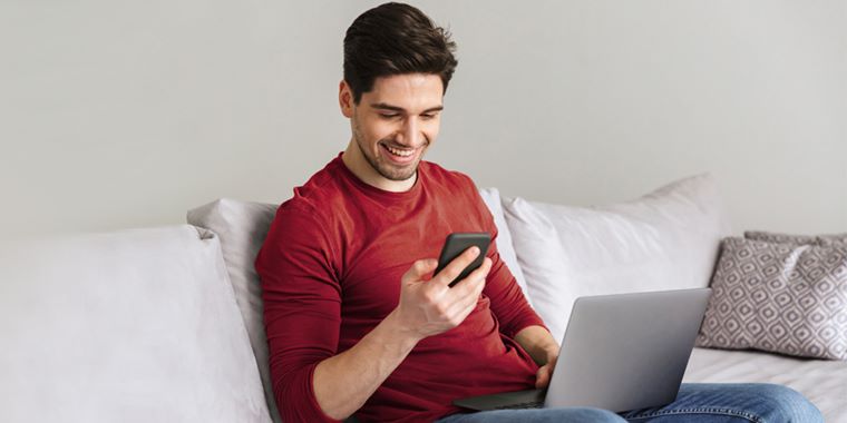 Moški v rdečem puloverju in modrih kavbojkah sedi na kavču. Gleda v pametni telefon, ki ga ima v levi roki, v naročju pa ima prenosni računalnik. V ozadju so bele stene, bele blazine in blazina z vzorcem.
