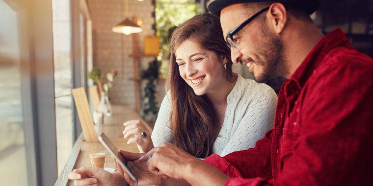 Moški v rdeči in ženska v beli srajci sedita ob oknu v kavarni. Gledata v zaslon pametnega telefona. Na mizi je skodelica kave.