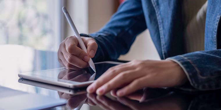 Oseba s pisalom piše na tablico, ki leži na stekleni mizi. Nahaja se v svetlem prostoru.