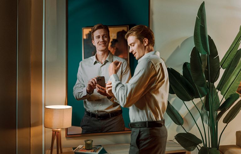 Dva moška stojita pred ogledalom. Levi drži telefon in gleda v ogledalo, desni si zapenja gumbe na srajci. V ozadju se vidi svetilka in velika zelena rastlina.