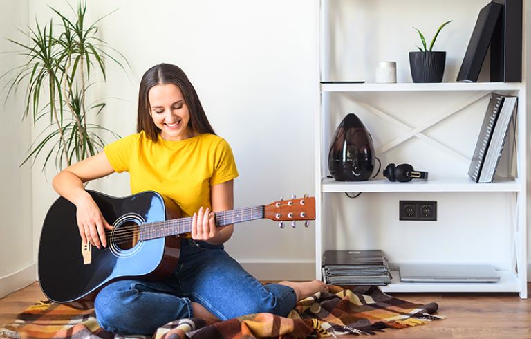 Dekle sedi na tleh in igra akustično kitaro. Oblečena je v rumeno majico in modre kavbojke. V ozadju je bela knjižna polica.