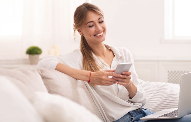 Mlada ženska sedi na kavču in gleda v pametni telefon. Oblečena je v belo srajco, ima rjave lase, ki so speti v čop. V ozadju je okno z zavesami ter majhna zelena rastlina, ki stoji na polici.