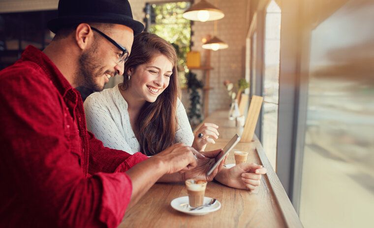 Moški v rdeči in ženska v beli srajci sedita ob oknu v kavarni. Gledata v zaslon pametnega telefona. Na mizi je skodelica kave.
