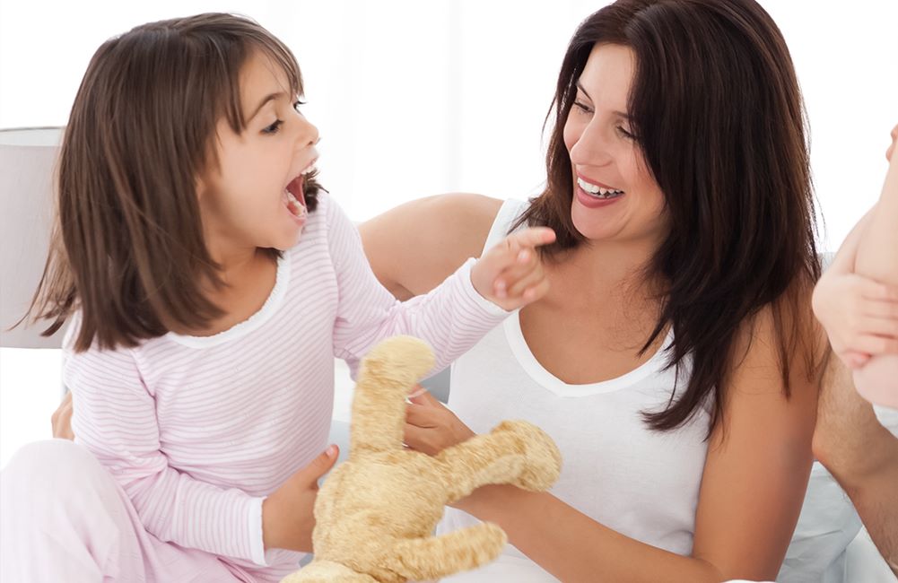 Punčka v roza pižami s plišastim medvedkom se igra z mamo, ki nosi belo majico in ima dolge črne lase.