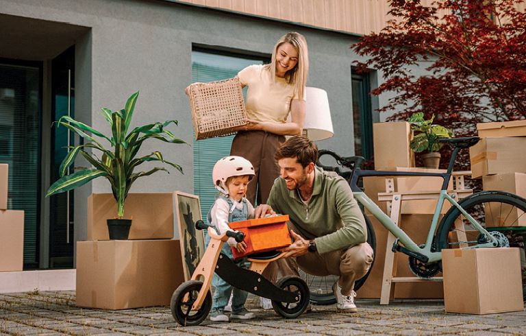 Družina se seli v nov dom. Stojijo pred hišo, obdani so s škatlami. Otrok in oče z oranžno škatlo čepita ob poganjalčku, mama stoji za njima.