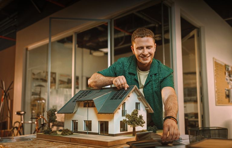 Moški v zeleni srajci izdeluje maketo hiše s sončnimi celicami in drevesi. V ozadju je prostor s steklenimi stenami.