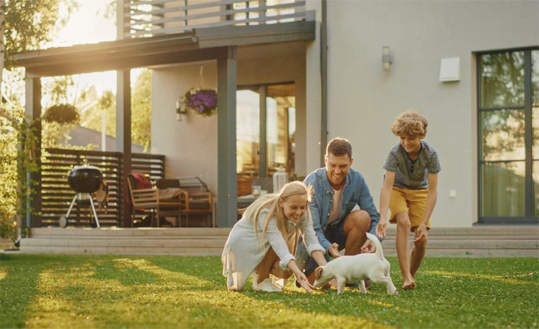 Tričlanska družina se z belim kužkom igra na travniku pred sodobno hišo z velikimi okni, balkonom in žarom v topli sončni svetlobi.