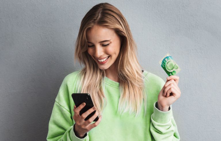 Ženska s svetlimi lasmi v zeleni majici gleda v pametni telefon in drži kreditno kartico. Ozadje na fotografiji je sivo.