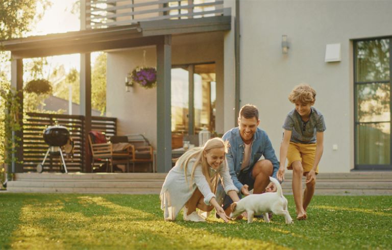 Tričlanska družina se z belim kužkom igra na travniku pred sodobno hišo z velikimi okni, balkonom in žarom v topli sončni svetlobi.
