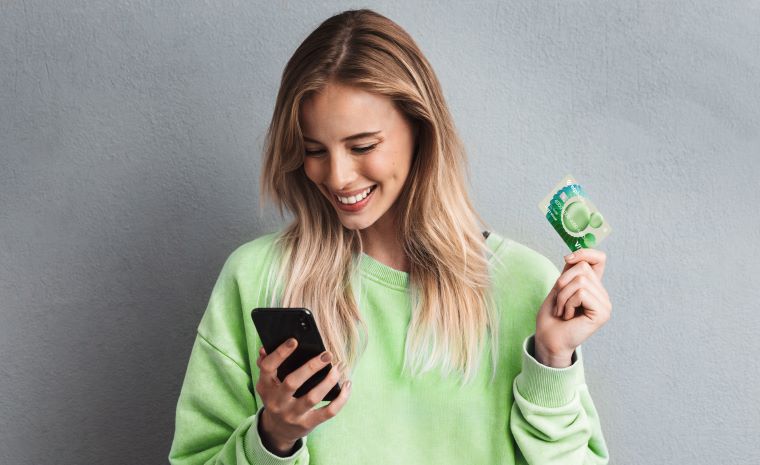 Ženska s svetlimi lasmi v zeleni majici gleda v pametni telefon in drži kreditno kartico. Ozadje na fotografiji je sivo.