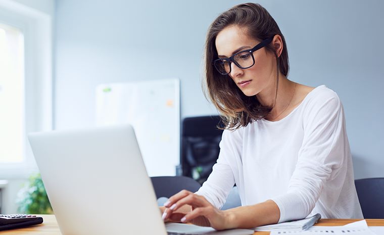 Ženska sedi za pisalno mizo in dela na prenosnem računalniku. Oblečena je v belo majico z dolgimi rokavi, ima srednje dolge rjave lase in nosi očala. Ženska sedi za pisalno mizo in dela na prenosnem računalniku. Oblečena je v belo majico z dolgimi rokavi, ima srednje dolge rjave lase in nosi očala.