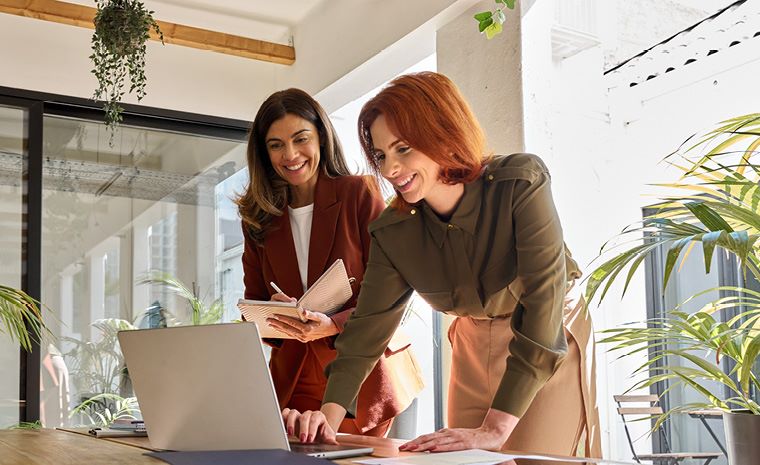 Ženski v pisarni stojita za mizo in gledata v računalnik. Na mizi so dokumenti, v ozadju pa steklene stene in rastline.